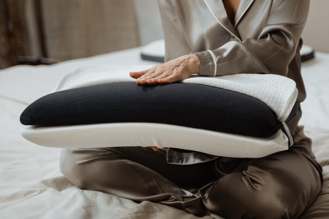 Eine Person in einem Seidenpyjama sitzt im Schneidersitz auf einem Bett und berührt ein schwarz-weißes Kissen, das aus mehreren Schichten besteht. Ihr Gesicht ist nicht zu sehen.