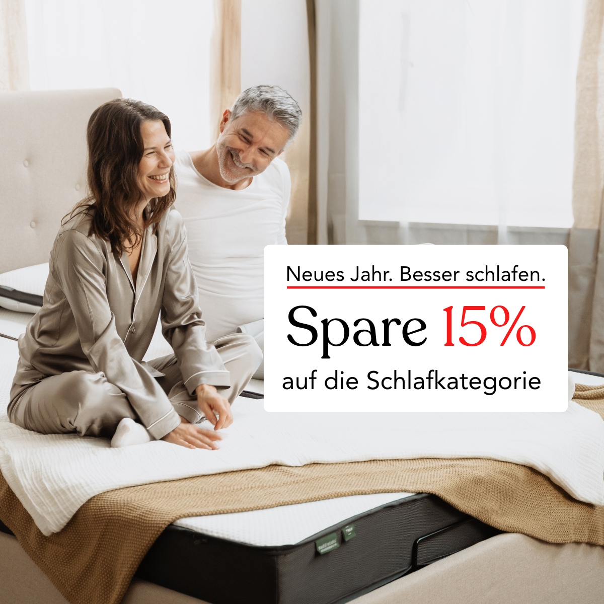 Ein lächelndes Paar sitzt auf einem ordentlich gemachten Bett in einem hellen Schlafzimmer. Textüberlagerung in Deutsch wirbt: Neues Jahr. Besser schlafen. Sparen Sie 15% in der Kategorie Schlaf.
