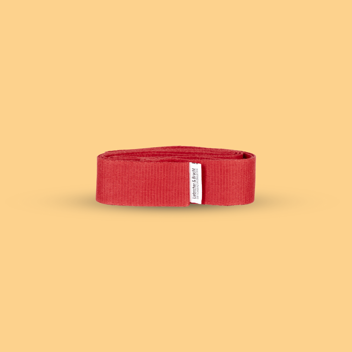 Die Liebscher & Bracht Übungs-Schlaufe, ein aufgerolltes rotes Widerstandsband mit einem kleinen weißen Etikett, ist ideal zum Üben und zur Steigerung der Beweglichkeit, abgebildet auf einem schlichten beigen Hintergrund.