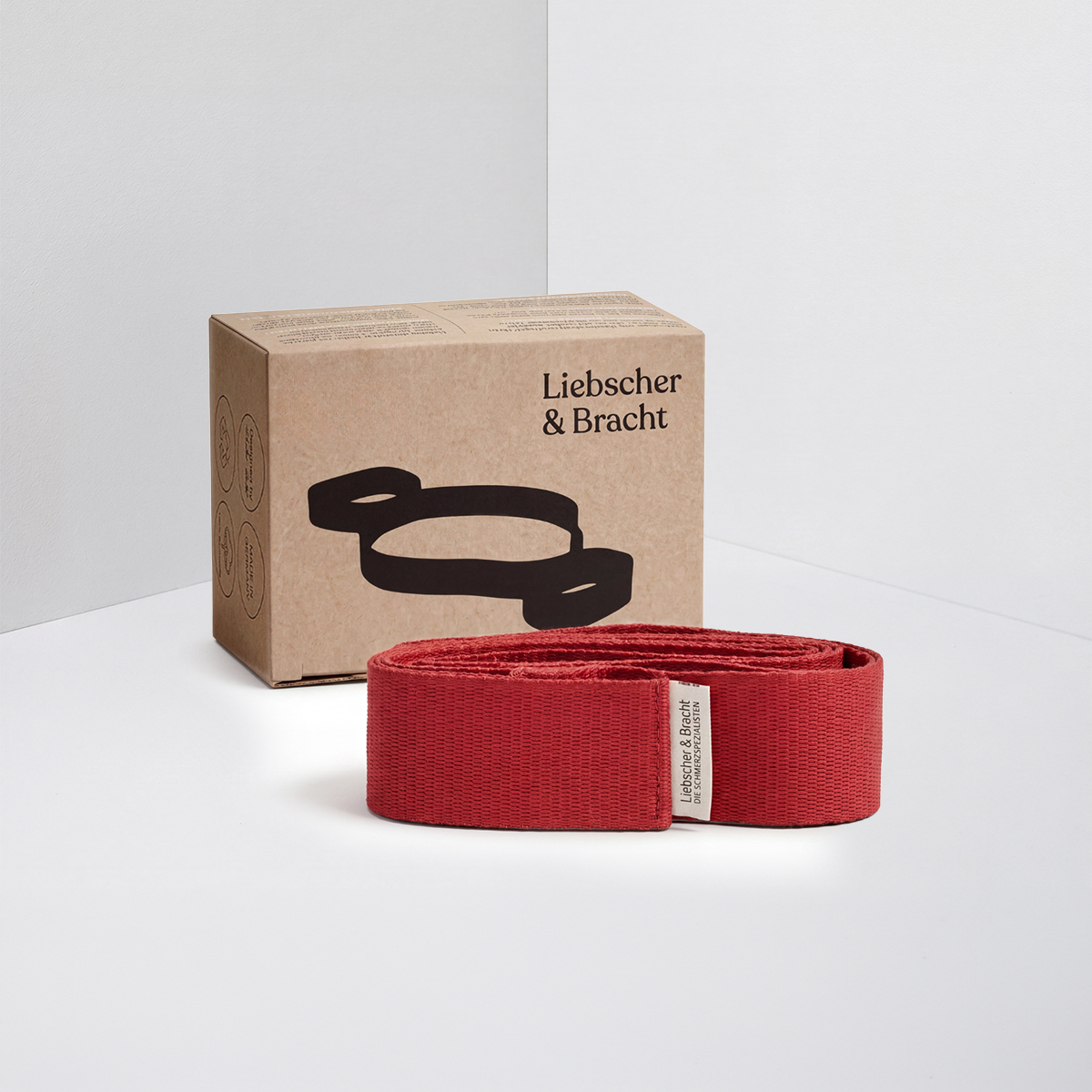 Ein rotes Übungsband der Marke „Übungs-Schlaufe“ von Liebscher & Bracht ist vor einer braunen Schachtel platziert, auf der der Markenname und eine schwarze Illustration zu sehen sind, vor einem weiß-hellgrauen Hintergrund.