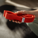 Eine Hand hält die „Liebscher & Bracht Übungs-Schlaufe“, ein aufgerolltes rotes Band mit Etikett, vor einem unscharfen Hintergrund mit einer strukturierten schwarzen Unterlage – ideal für gezieltes Üben und mehr Beweglichkeit bei Dehnübungen.