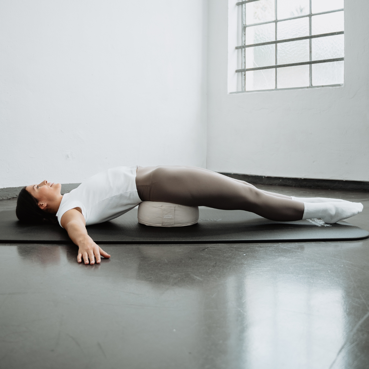 Eine Person in Sportkleidung liegt mit ausgestreckten Armen und angewinkelten Knien auf einer Yogamatte über dem „Liebscher & Bracht Übungskissen-Set“ in einem hellen, minimalistisch eingerichteten Raum mit einem großen Fenster.