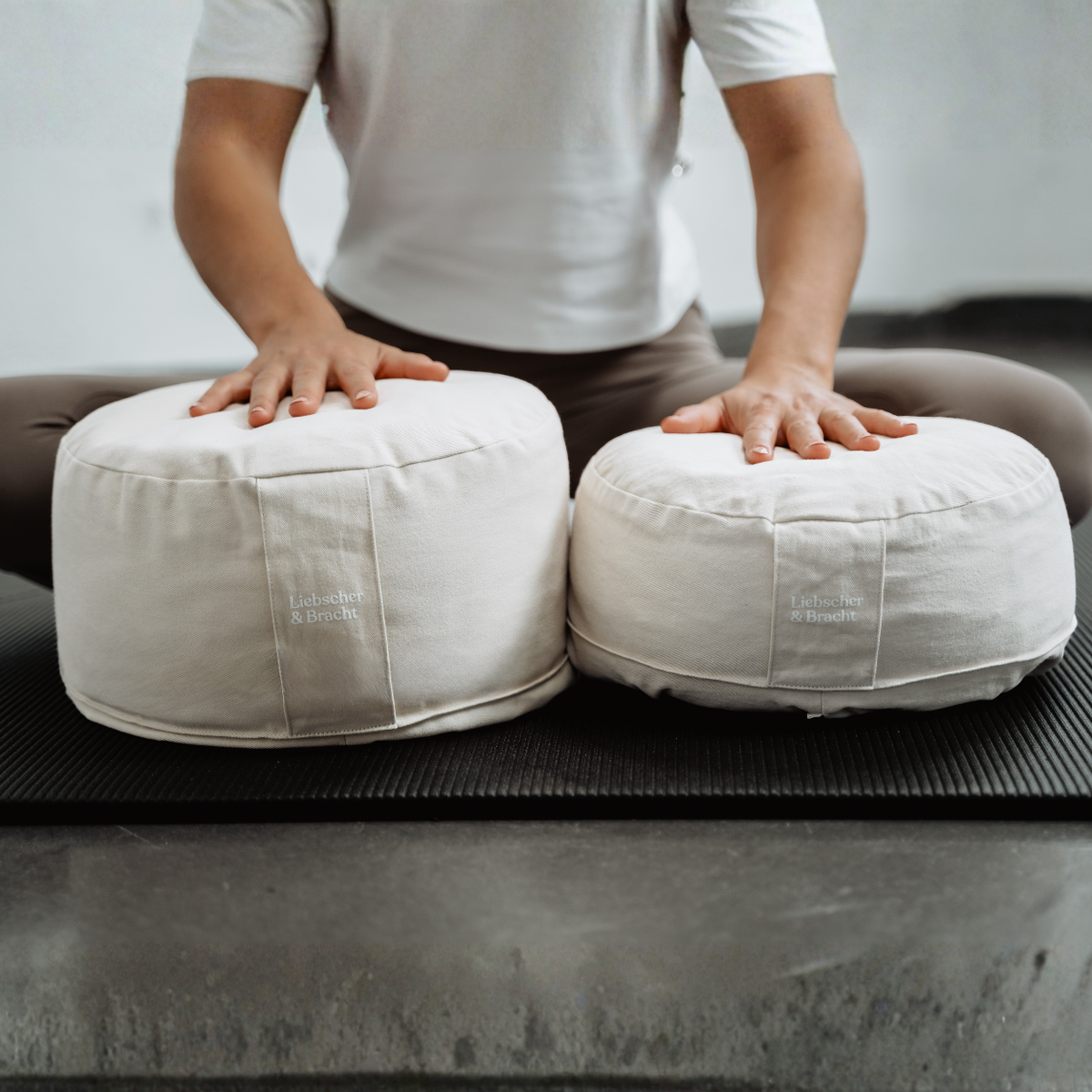 Eine Person in einem weißen Hemd sitzt mit gekreuzten Beinen auf einer Yogamatte und stützt sich mit den Händen auf zwei weiße Kissen des „Liebscher & Bracht Übungskissen-Sets“ ab, die vor ihr liegen.