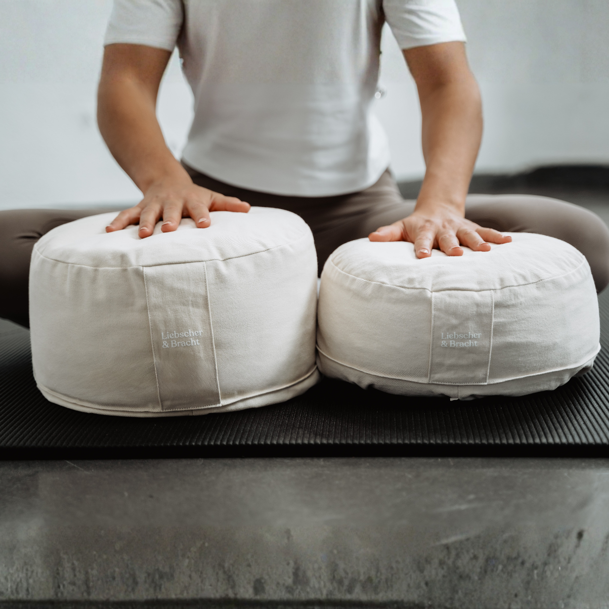 Eine Person in einem weißen Hemd sitzt mit gekreuzten Beinen auf einer Yogamatte und stützt sich mit den Händen auf zwei weiße Kissen des „Liebscher & Bracht Übungskissen-Sets“ ab, die vor ihr liegen.