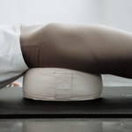 Eine Person in braunen Leggings und einem weißen Shirt liegt auf einer Yogamatte, wobei ihre Hüften vom Liebscher & Bracht Übungskissen-Set gestützt werden.
