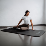 Eine Frau in einem weißen Shirt und braunen Leggings macht Dehnübungen auf der rutschfesten Rolands Übungsmatte von Liebscher & Bracht in einem minimalistisch eingerichteten Raum – so wird ihr Training stilvoll und gelenkschonend.