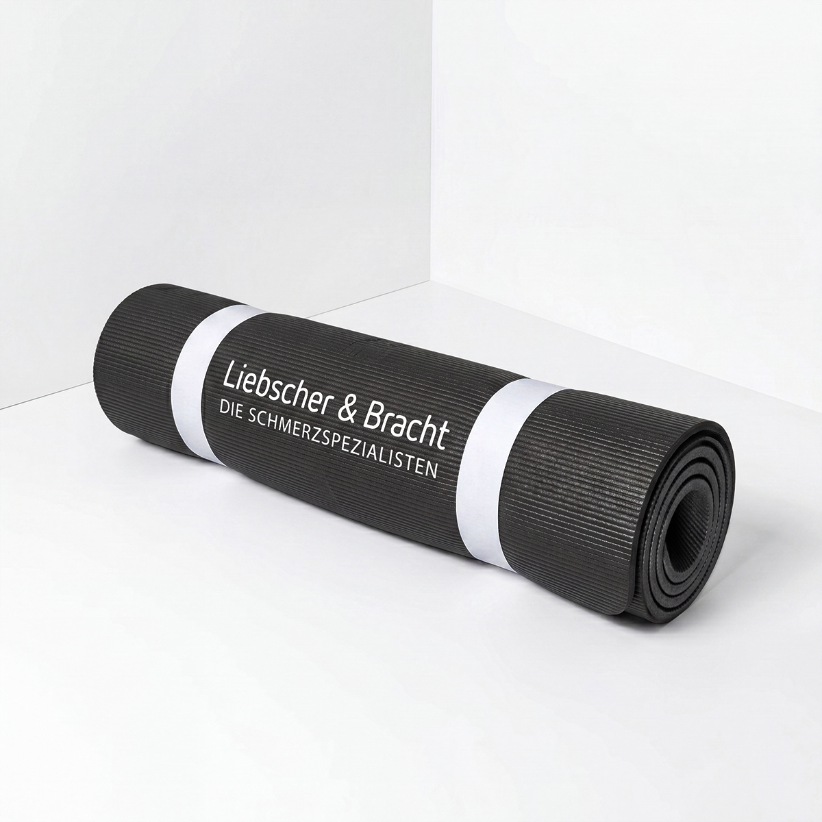 Eine schwarze, rutschfeste Rolands-Übungsmatte von Liebscher & Bracht mit weißem Logo ist aufgerollt und liegt in einer hellen Ecke.