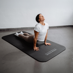 Eine Frau in Sportkleidung macht drinnen Yoga und führt die „Upward-Facing Dog“-Pose auf einer „Rolands“-Übungsmatte von Liebscher & Bracht aus, die auf einem grauen Boden liegt.