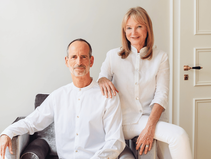 Ein Mann und eine Frau in weißen Kleidern posieren in einem Innenraum. Der Mann sitzt auf einem grauen Stuhl, während die Frau neben ihm steht und ihre Hand auf seiner Schulter ruht. Beide lächeln und der Hintergrund ist minimal und sanft beleuchtet.