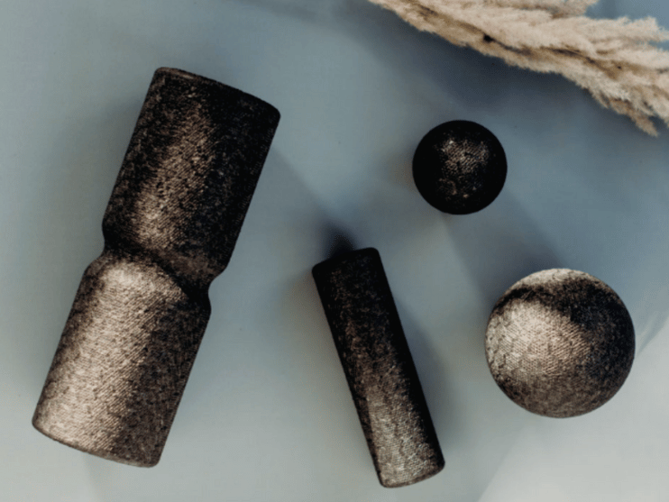 Vier schwarze, strukturierte Massagewerkzeuge - zwei zylindrische Rollen und zwei Massagebälle - sind auf einer hellblauen Fläche angeordnet, mit beigem Pampasgras in der oberen rechten Ecke.