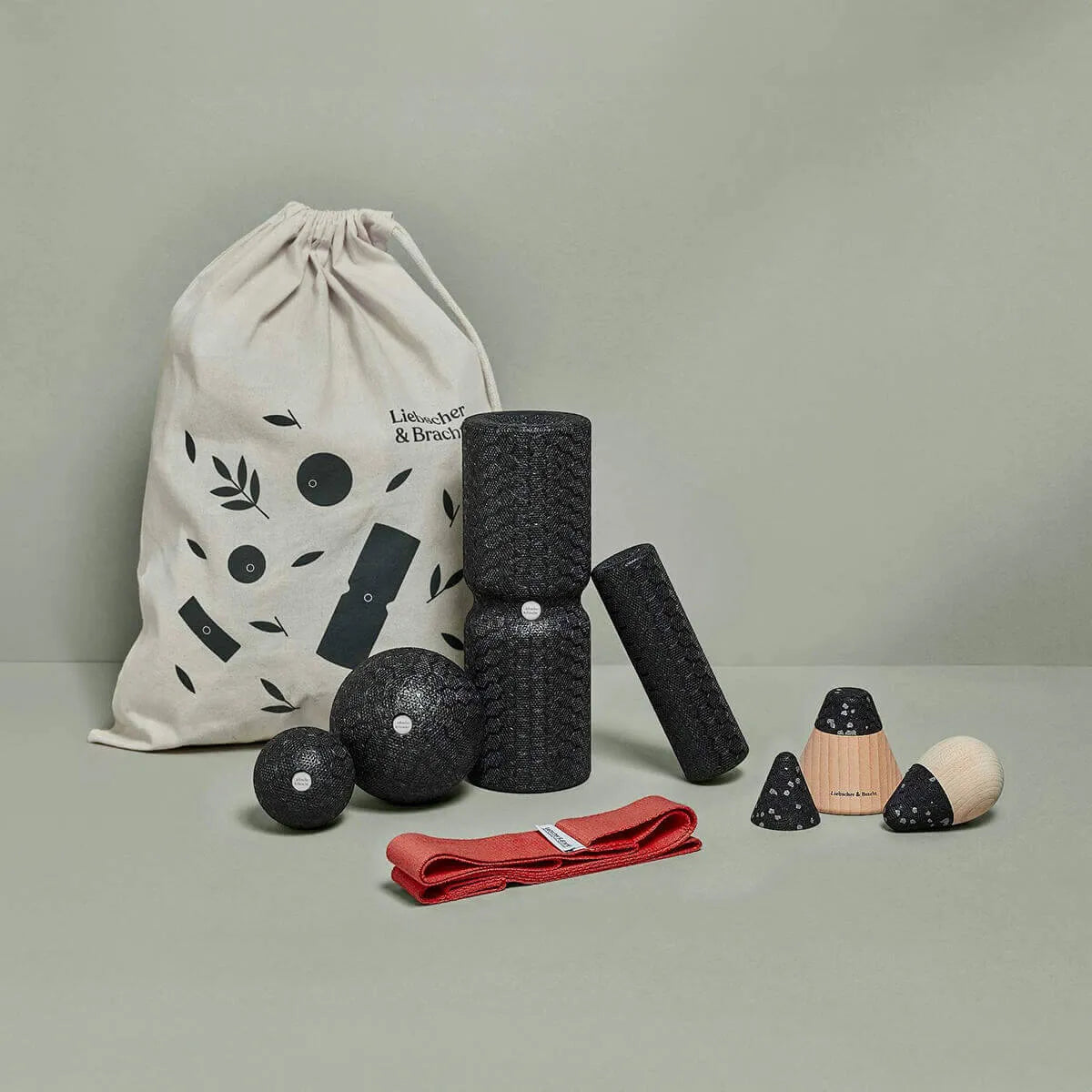 Das Angebots-Paket von Liebscher & Bracht mit schwarzen, texturierten Foam Rollern, Bällen, Kegeln, roter Übungsschlaufe und beigem Beutel mit Muster liegt auf hellgrauem Untergrund vor grauem Hintergrund.