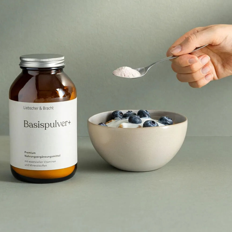 Eine Hand hält einen Löffel mit Pulver über einer Schale mit Joghurt und Blaubeeren. Neben der Schale steht ein braunes Glasgefäß mit der Aufschrift Basispulver+ von Liebscher & Bracht. Die Szene ist vor einem schlichten Hintergrund dargestellt.