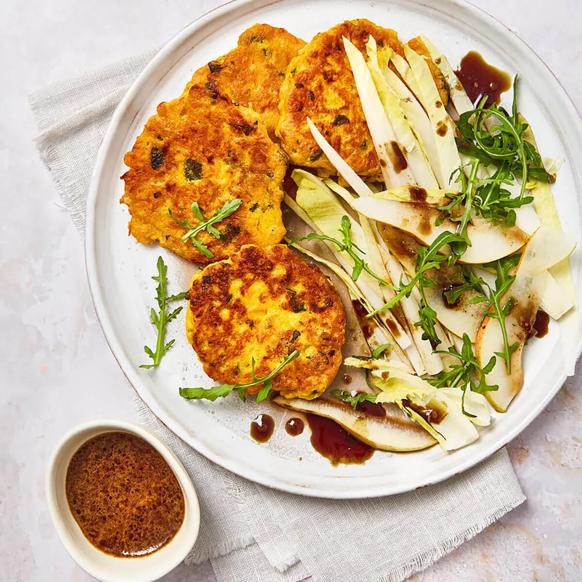 Auf einem weißen Teller liegen drei goldbraune Krapfen neben einem Salat aus gehobeltem Fenchel, Birne und Rucola - genau richtig für gesund abnehmen mit Abnehmen garantiert von Liebscher & Bracht. Auf einer leichten Stoffserviette ruht ein Schälchen mit extra Dressing.