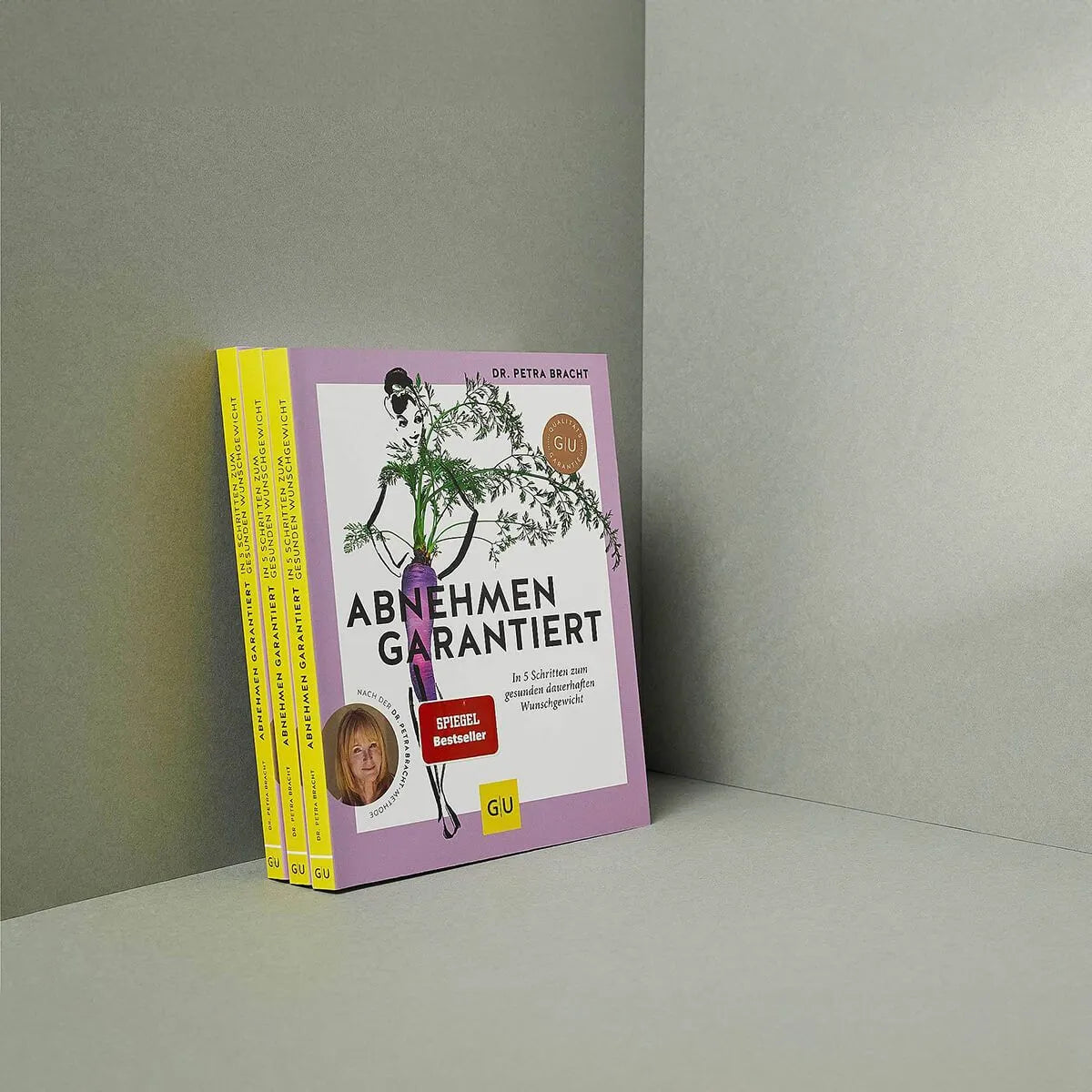 Das Buch "Abnehmen garantiert" von Liebscher & Bracht mit lila Einband und botanischer Illustration steht aufrecht neben zwei gestapelten Exemplaren vor minimalem grauen Hintergrund. Es bietet Tipps für gesundes Abnehmen und pflanzliche Rezepte.