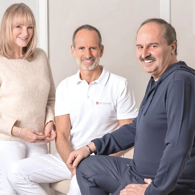 Drei Erwachsene lächeln in die Kamera: eine Frau in einem beigen Pullover, ein Mann in einem weißen Polohemd und ein weiterer Mann in einer marineblauen Jacke, die alle in einem Haus vor einem hellen Hintergrund sitzen.