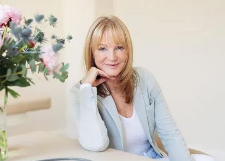 Eine Frau mit blondem Haar sitzt an einem hellen Tisch, lächelt und stützt ihren Kopf auf ihre Hand. Eine Vase mit rosa Blumen und ein Stethoskop stehen auf dem Tisch in einem hellen, minimalistischen Raum.