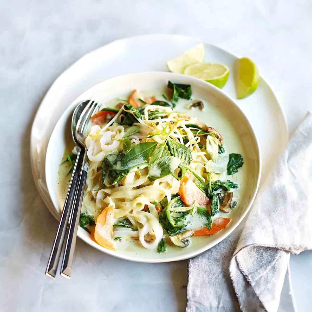 Eine weiße Schale mit cremiger Gemüsenudelsuppe, inspiriert von veganen Rezepten aus dem "Intervallfasten Kochbuch" von Liebscher & Bracht, wird mit frischen Kräutern und Limettenspalten garniert und mit Gabel, Löffel und einer leichten Serviette serviert.