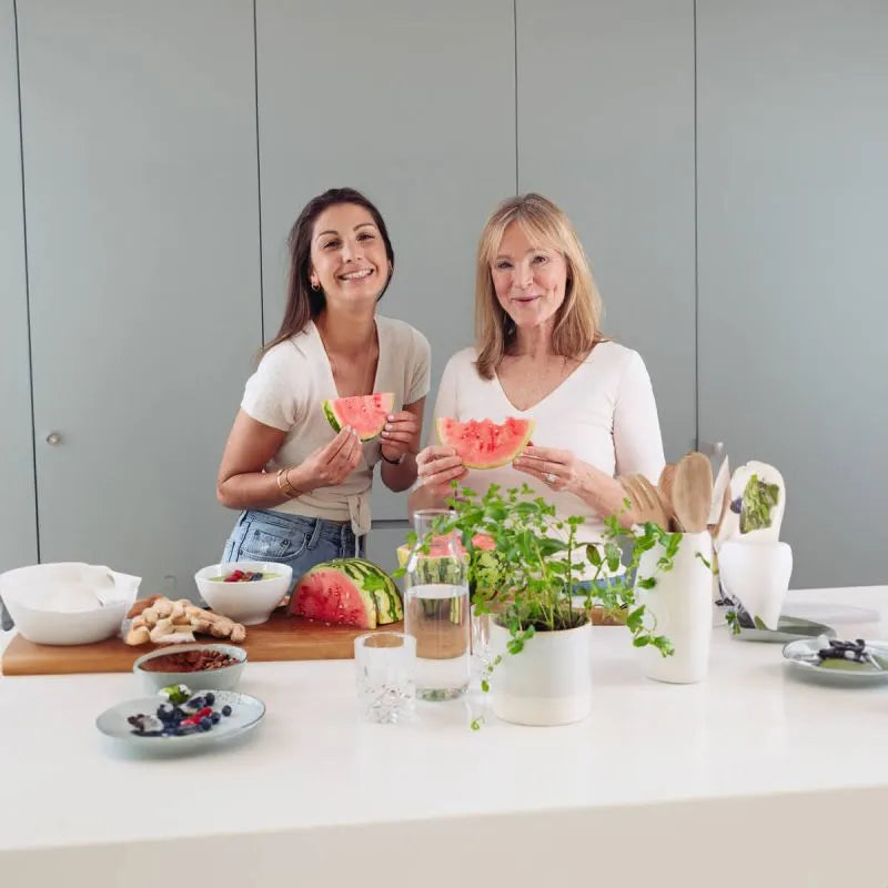 Zwei Frauen stehen in einer modernen Küche, lächeln und halten Scheiben von Wassermelonen in der Hand. Auf der Theke vor ihnen stehen Teller mit Beeren, eine Pflanze und verschiedene Küchenutensilien. Beide tragen helle Oberteile.