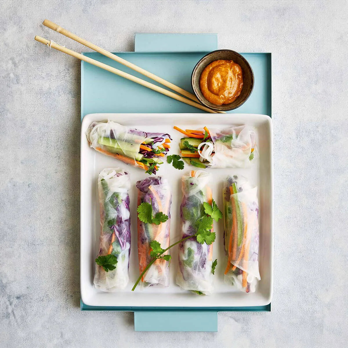 Ein weißes Tablett mit fünf frischen Gemüse-Frühlingsrollen, Dip-Sauce und Essstäbchen auf blauem Hintergrund - ideal für Rezepte aus dem Intervallfasten Kochbuch von Liebscher & Bracht.