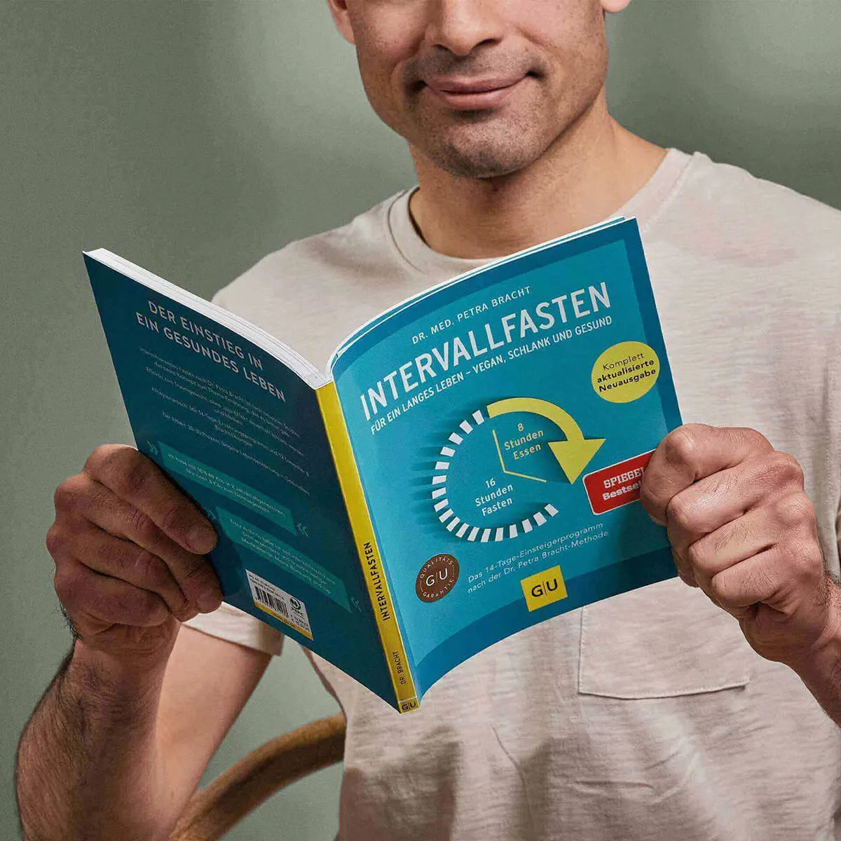 Ein Mann in einem beigen Hemd hält und liest "Intervallfasten" von Liebscher & Bracht, einen deutschen Ratgeber für gesunde Ernährung und dauerhaftes Abnehmen. Der blaue Einband hat gelbe und weiße Akzente und zeigt ein Fastendiagramm.