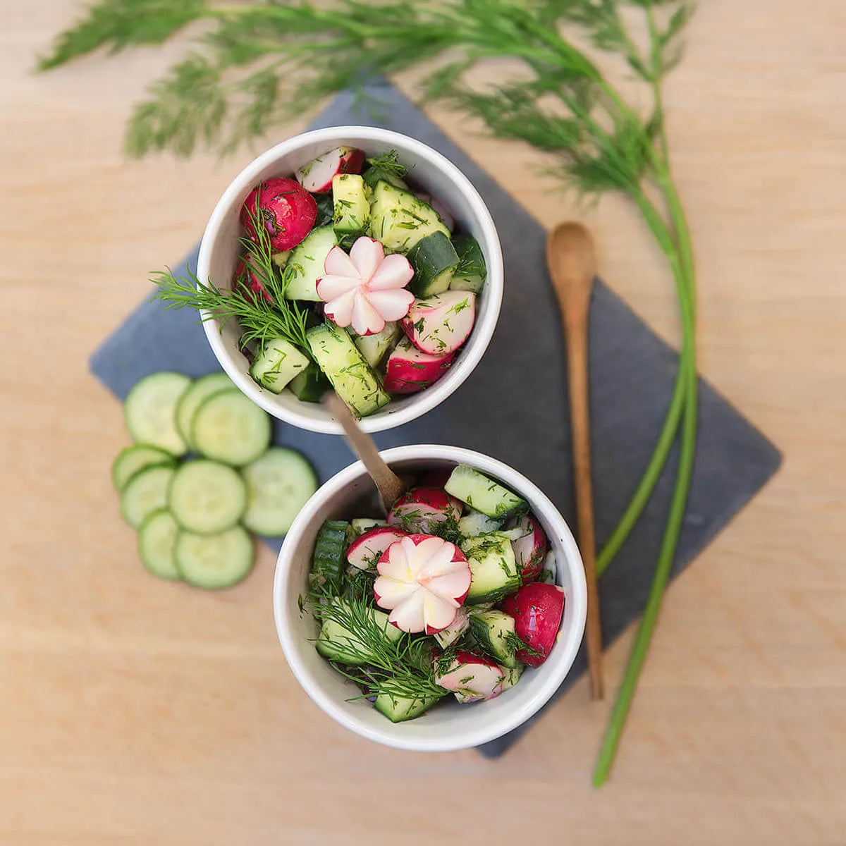 Zwei Schalen Gurken- und Rettichsalat mit Dill, perfekt für pflanzliche Rezepte, gestylt auf einer Schiefertafel mit Gurkenscheiben und Dill. Ausgestattet mit Intervallfasten von Liebscher & Bracht auf einer hellen Holzoberfläche.