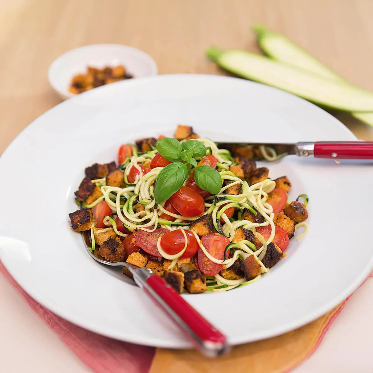 Ein Teller mit Zucchininudeln, Kirschtomaten, Basilikum und Croutons ist ein Beispiel für gesunde Ernährung - ideal für die Verwendung von Intervallfasten von Liebscher & Bracht. Gabel und Messer liegen auf dem Teller, im Hintergrund sind Zucchinischeiben und Croutons zu sehen.