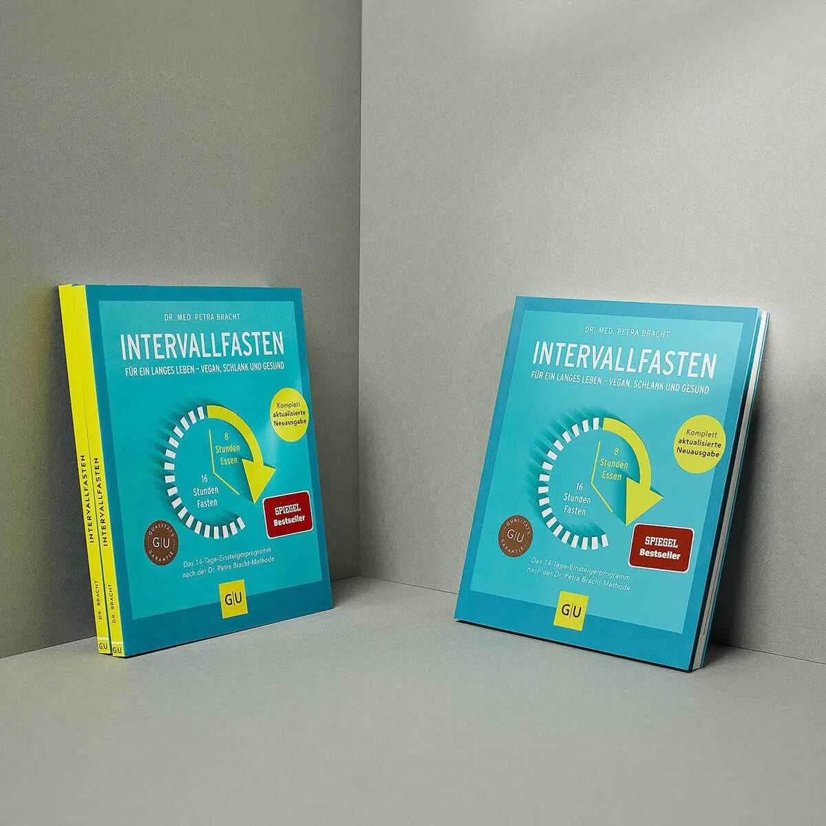 Zwei Exemplare des Buches Intervallfasten von Liebscher & Bracht mit tealfarbenem Einband und Uhrendesign sind auf hellgrauem Hintergrund abgebildet - eines hochkant, eines flach - ideal für Tipps zur gesunden Ernährung und zum dauerhaften Abnehmen.