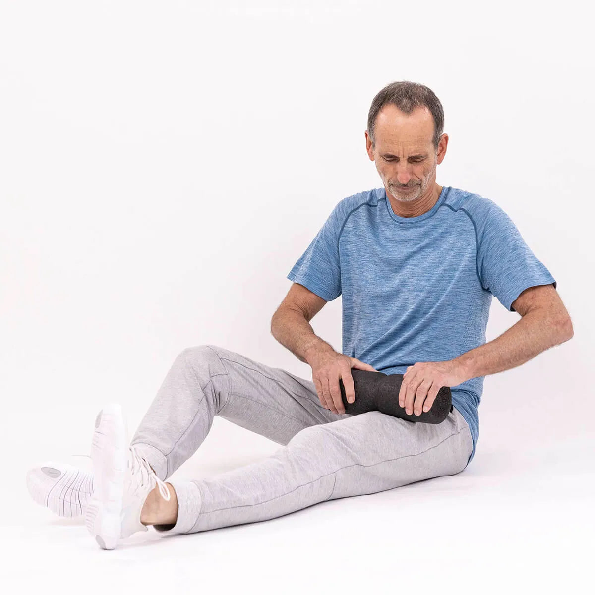 Ein Mann mittleren Alters in blauem Hemd und grauer Jogginghose sitzt mit einer Schaumstoffrolle auf dem Boden und wendet die Liebscher & Bracht-Methode "Ischias-Schmerzen selbst behandeln" zur Linderung und effektiven Schmerztherapie bei Ischialgie an.