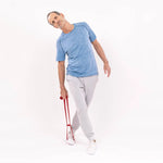 Ein Mann in blauem Hemd und grauer Hose demonstriert eine Dehnung mit einem roten Widerstandsband, wie sie in "Ischias-Schmerzen selbst behandeln" von Liebscher & Bracht zur ISG-Schmerzlinderung empfohlen wird. Der Hintergrund ist einfarbig weiß.