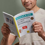 Ein Mann im hellen Hemd lächelt, während er das aufgeschlagene Buch "Ischias-Schmerzen selbst behandeln" von Liebscher & Bracht in der Hand hält, das Methoden zur Selbstbehandlung von Ischias- und Iliosakralgelenkschmerzen enthält.