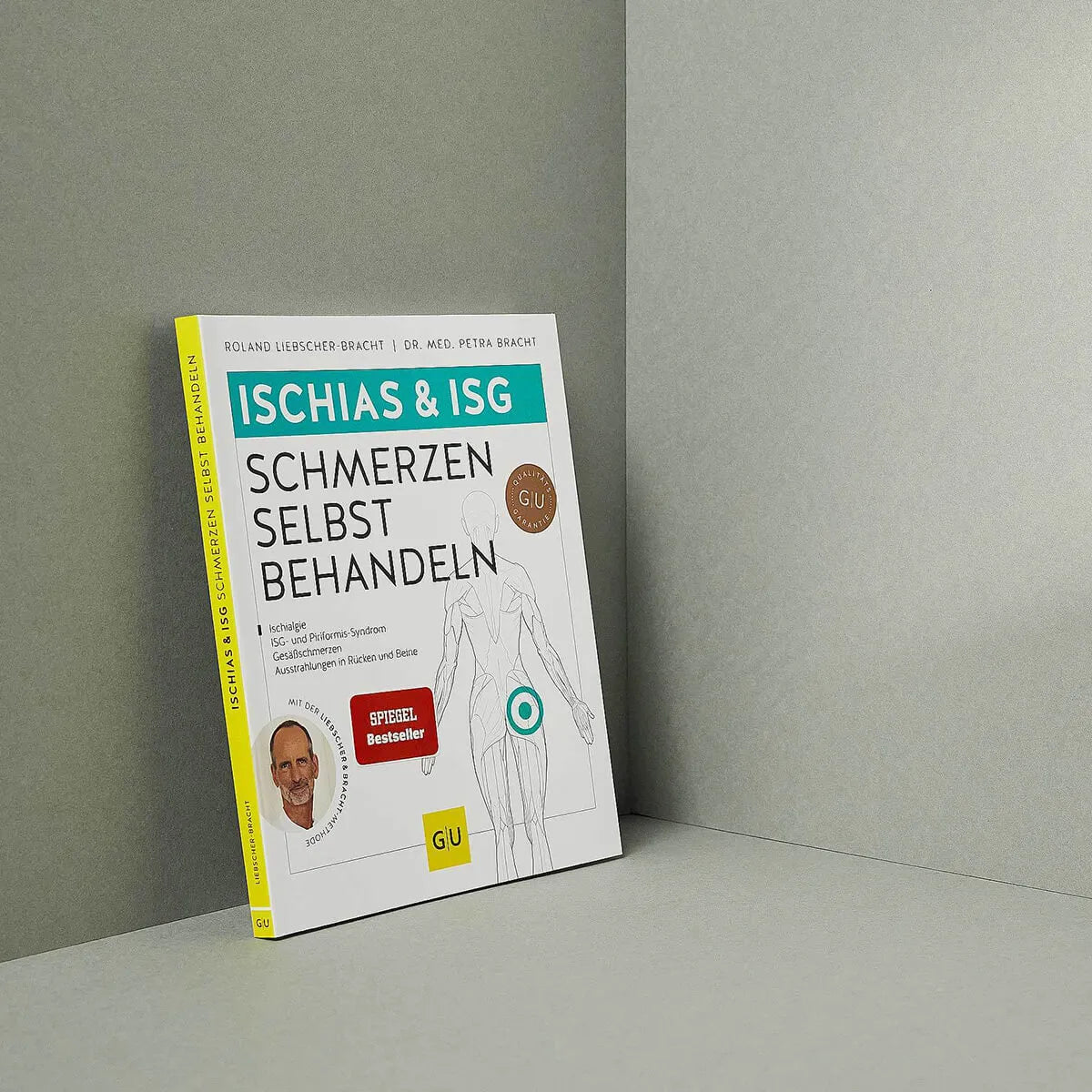 Das deutsche Buch Ischias-Schmerzen selbst behandeln von Liebscher & Bracht steht aufrecht an einer grauen Wand und zeigt Methoden der Schmerztherapie, ein rundes Abzeichen und ein kleines Foto eines lächelnden Mannes in der unteren linken Ecke.