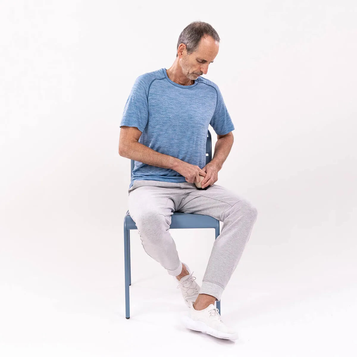 Ein Mann mit kurzen grauen Haaren sitzt auf einem blauen Stuhl, zieht ein blaues Hemd hoch und verwaltet Ischias-Schmerzen. Er trägt eine hellgraue Jogginghose und weiße Turnschuhe. Die Szene wirbt für "Ischias-Schmerzen selbst behandeln" von Liebscher & Bracht.