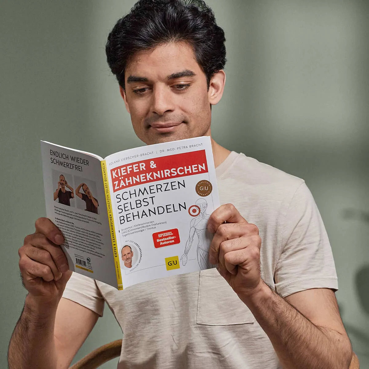 Ein dunkelhaariger Mann in einem hellen Hemd sitzt im Haus und liest das Buch "Kieferschmerzen & Zähneknirschen selbst behandeln" von Liebscher & Bracht, das ein wirksames Übungsprogramm gegen Kieferschmerzen und Zähneknirschen bietet.