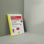 Ein Stapel "Kieferschmerzen & Zähneknirschen selbst behandeln" von Liebscher & Bracht steht aufrecht vor einem grauen Hintergrund. Der Umschlag zeigt gelbe und rote Schrift, ein kleines Autorenfoto und ein Übungsprogramm für Kieferschmerzen-Relief.