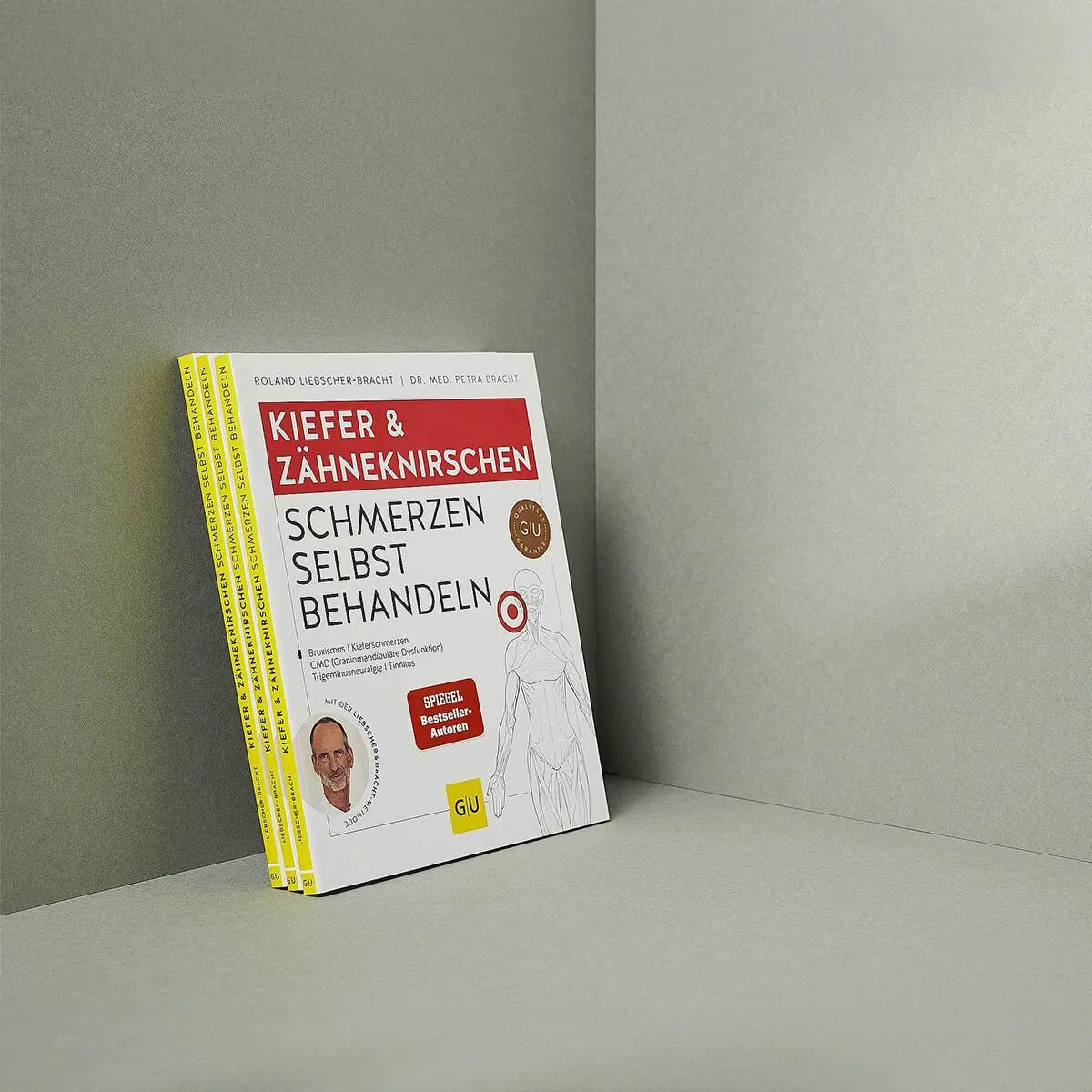 Ein Stapel "Kieferschmerzen & Zähneknirschen selbst behandeln" von Liebscher & Bracht steht aufrecht vor einem grauen Hintergrund. Der Umschlag zeigt gelbe und rote Schrift, ein kleines Autorenfoto und ein Übungsprogramm für Kieferschmerzen-Relief.