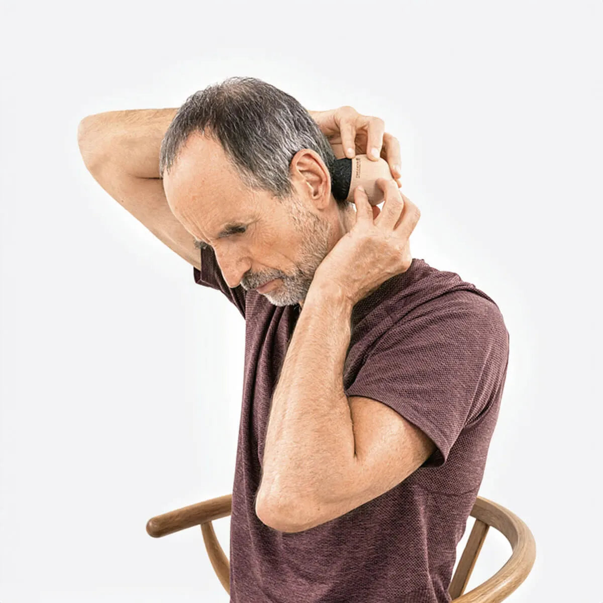 Ein Mann mit kurzen grauen Haaren und kastanienbraunem Hemd sitzt auf einem Holzstuhl und greift sich in den Nacken, um Liebscher & Brachts "Kieferschmerzen & Zähneknirschen selbst behandeln" anzuwenden, möglicherweise als Teil eines Übungsprogramms zur Linderung von Kieferschmerzen.
