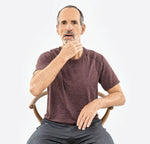 Ein Mann mittleren Alters mit kurzen Haaren und Bart demonstriert eine Übung aus "Kieferschmerzen & Zähneknirschen selbst behandeln" von Liebscher & Bracht, indem er im Sitzen seinen Hals berührt. Er trägt ein kastanienbraunes T-Shirt und eine graue Hose.