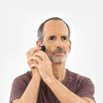 Ein bärtiger Mann in einem kastanienbraunen Hemd demonstriert vor einem schlichten hellen Hintergrund das Liebscher & Bracht-Werkzeug "Kieferschmerzen & Zähneknirschen selbst behandeln" auf seiner Wange.