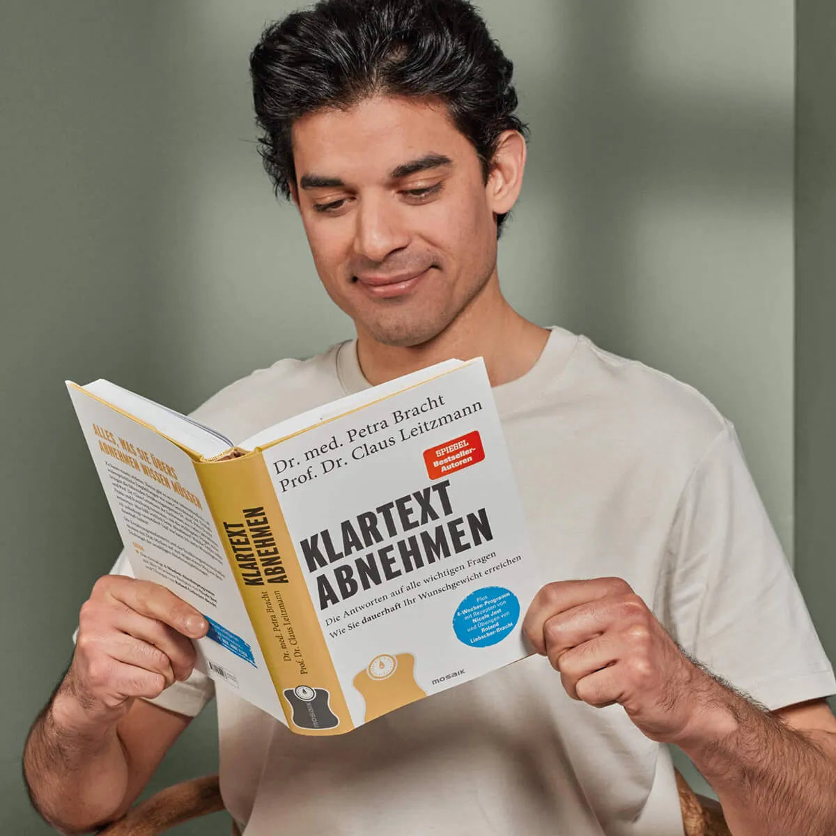 Ein dunkelhaariger Mann in einem hellen T-Shirt lächelt, während er "Klartext Abnehmen" von Liebscher & Bracht, ein deutsches Buch über ein 4-Wochen-Programm zum Erreichen Ihres Wunschgewichts, vor einem schlichten hellgrünen Hintergrund liest.