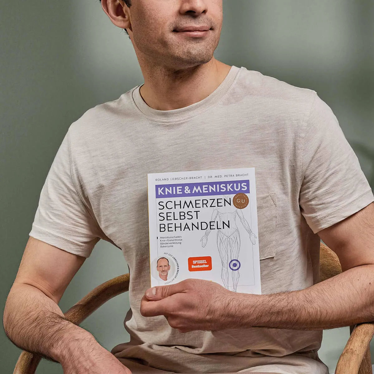 Ein Mann in einem beigen T-Shirt sitzt auf einem Stuhl und hält das Buch "Knieschmerzen selbst behandeln" von Liebscher & Bracht in der Hand, das auf dem Einband eine Teilabbildung eines Beins und ein Autorenfoto zeigt.