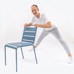 Ein älterer Mann in weißem Hemd und grauer Hose demonstriert eine Seitendehnung aus "Knieschmerzen selbst behandeln" von Liebscher & Bracht und hält dabei die Lehne eines blauen Stuhls vor einem schlichten weißen Hintergrund.