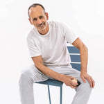 Ein älterer Mann in weißem T-Shirt und grauer Hose massiert seinen Oberschenkel mit einem Massageball nach dem Programm "Knieschmerzen selbst behandeln" von Liebscher & Bracht, um Knieschmerzen zu lindern. Er sitzt lächelnd auf einem blauen Stuhl vor einem schlichten weißen Hintergrund.