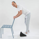 Ein Mann in weißem T-Shirt und hellgrauer Jogginghose streckt seine Wade auf einem Schrägbrett und stützt sich dabei auf einen hellblauen Stuhl - Teil des Programms "Knieschmerzen selbst behandeln" von Liebscher & Bracht - vor weißem Hintergrund.