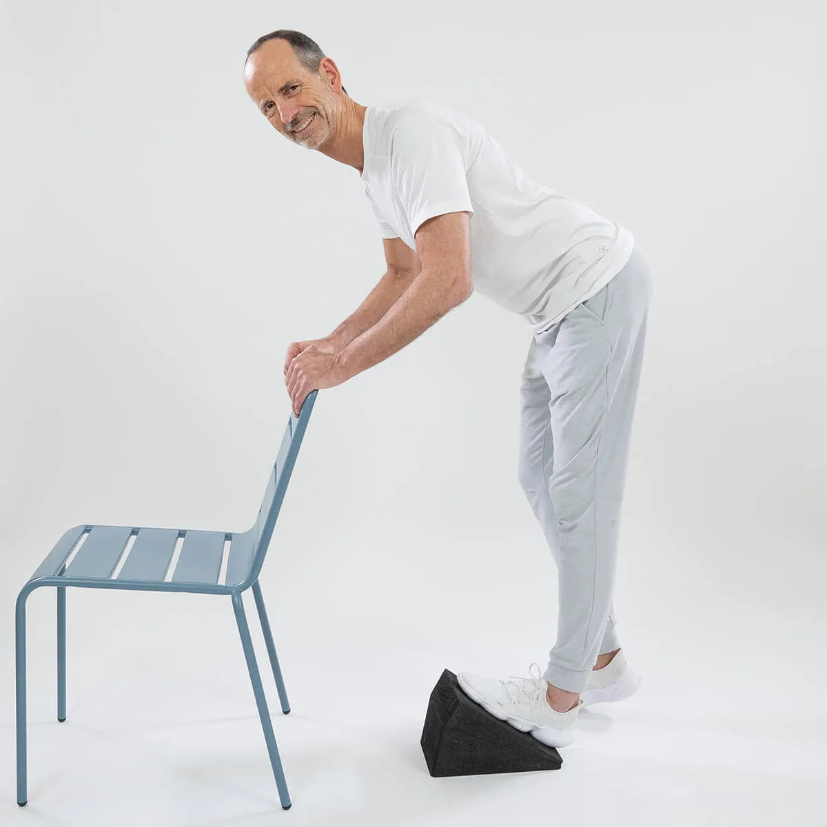 Ein Mann in weißem T-Shirt und hellgrauer Jogginghose streckt seine Wade auf einem Schrägbrett und stützt sich dabei auf einen hellblauen Stuhl - Teil des Programms "Knieschmerzen selbst behandeln" von Liebscher & Bracht - vor weißem Hintergrund.