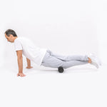 Ein Mann in Sportkleidung zeigt ein Brett mit einer Schaumstoffrolle unter den Oberschenkeln und demonstriert das Programm "Knieschmerzen selbst behandeln" von Liebscher & Bracht vor einem schlichten weißen Hintergrund.