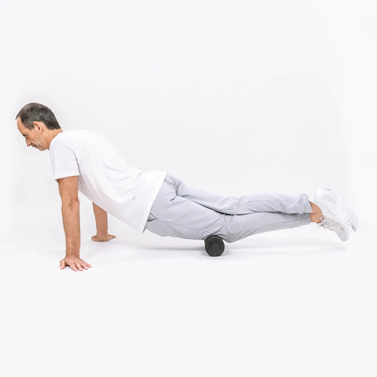 Ein Mann in Sportkleidung zeigt ein Brett mit einer Schaumstoffrolle unter den Oberschenkeln und demonstriert das Programm "Knieschmerzen selbst behandeln" von Liebscher & Bracht vor einem schlichten weißen Hintergrund.