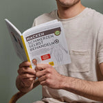 Ein Mann in einem beigen Hemd hält das aufgeschlagene Buch "Nackenschmerzen selbst behandeln" von Liebscher & Bracht in der Hand, das anatomische Abbildungen, ein forschungsbasiertes Nackenschmerzprogramm und das Foto eines Mannes auf dem deutschen Einband zeigt.