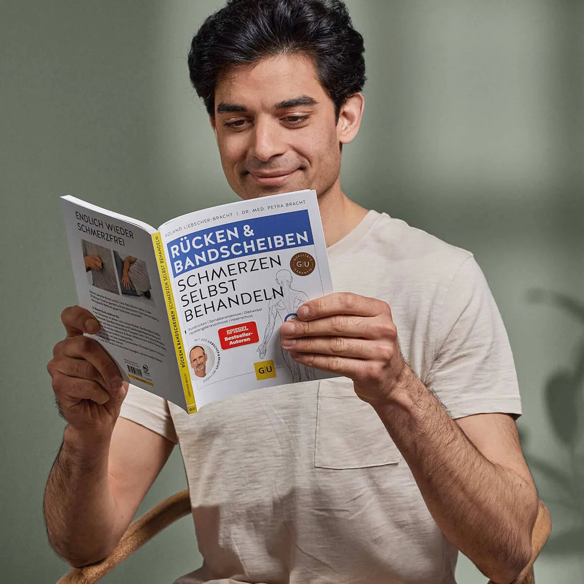 Ein dunkelhaariger Mann in einem hellbeigen T-Shirt sitzt da und lächelt, während er "Rücken- & Bandscheibenschmerzen selbst behandeln" von Liebscher & Bracht liest. Der Hintergrund ist schlicht und sanft beleuchtet.
