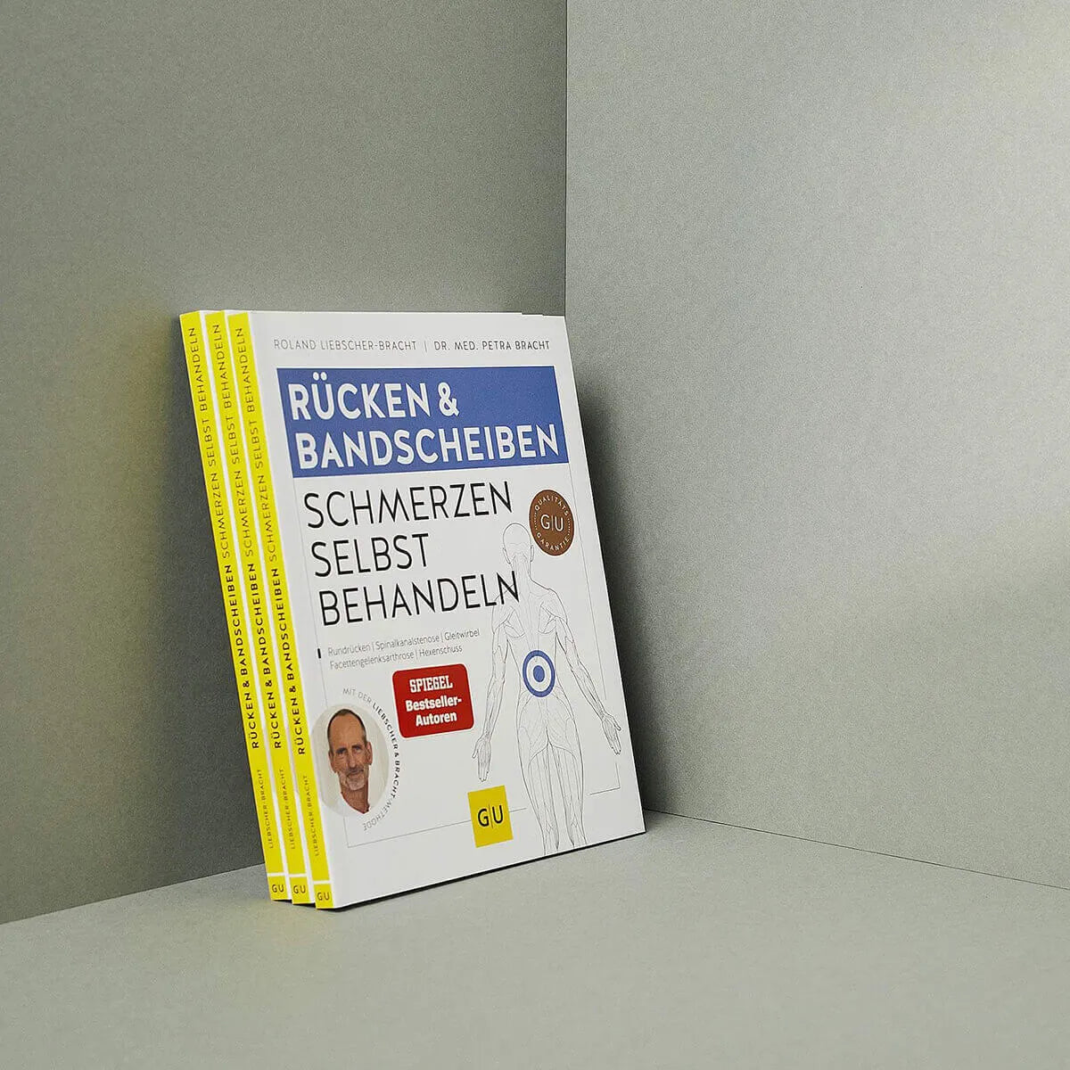 Vier Exemplare von "Rücken- & Bandscheibenschmerzen selbst behandeln" von Liebscher & Bracht, mit weißem Einband und gelbem Rücken, stehen aufrecht in einer schlichten grauen Ecke.