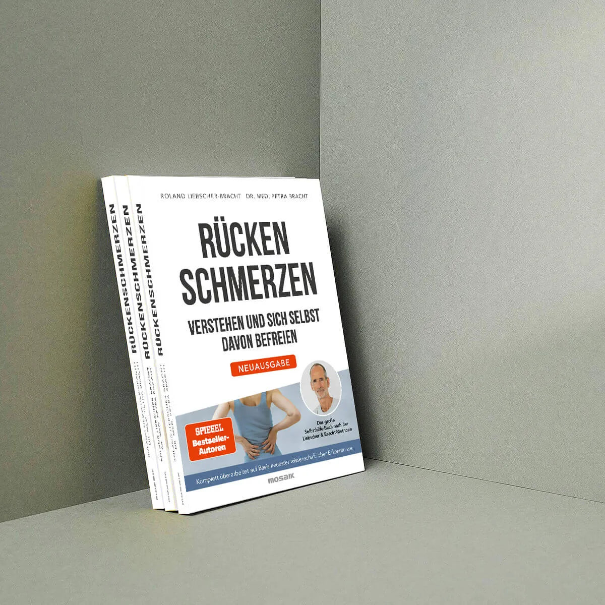 Drei Exemplare von "Rückenschmerzen verstehen und sich selbst davon befreien" von Liebscher & Bracht stehen aufrecht vor einer grauen Wand. Das Cover zeigt eine Person, die sich vor Schmerzen den unteren Rücken hält.